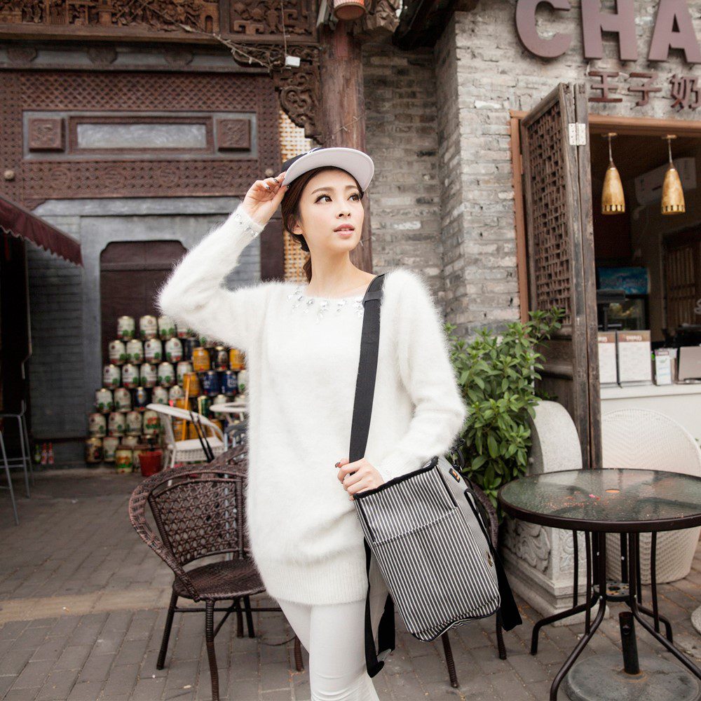 Jeune femme en pull blanc moelleux, sac rayé en bandoulière sur la hanche, debout devant une boutique en bois