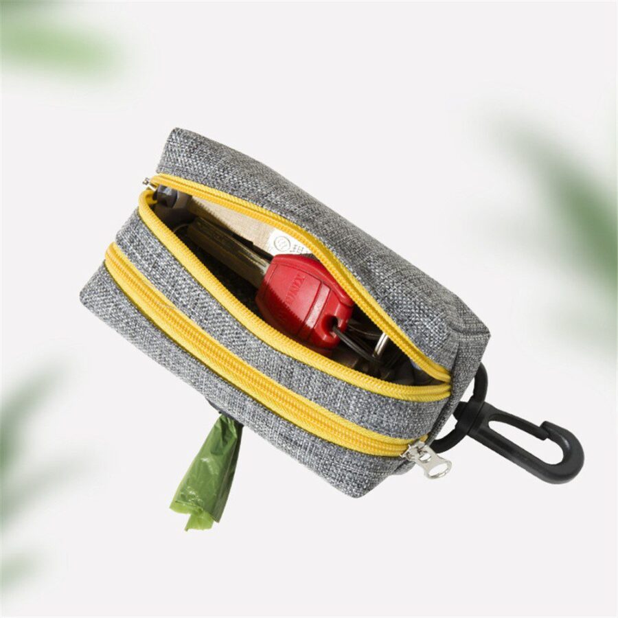 Petit distributeur gris à double fermeture jaune, ouvert sur une clé rouge, avec un sac vert qui dépasse et mousqueton noir