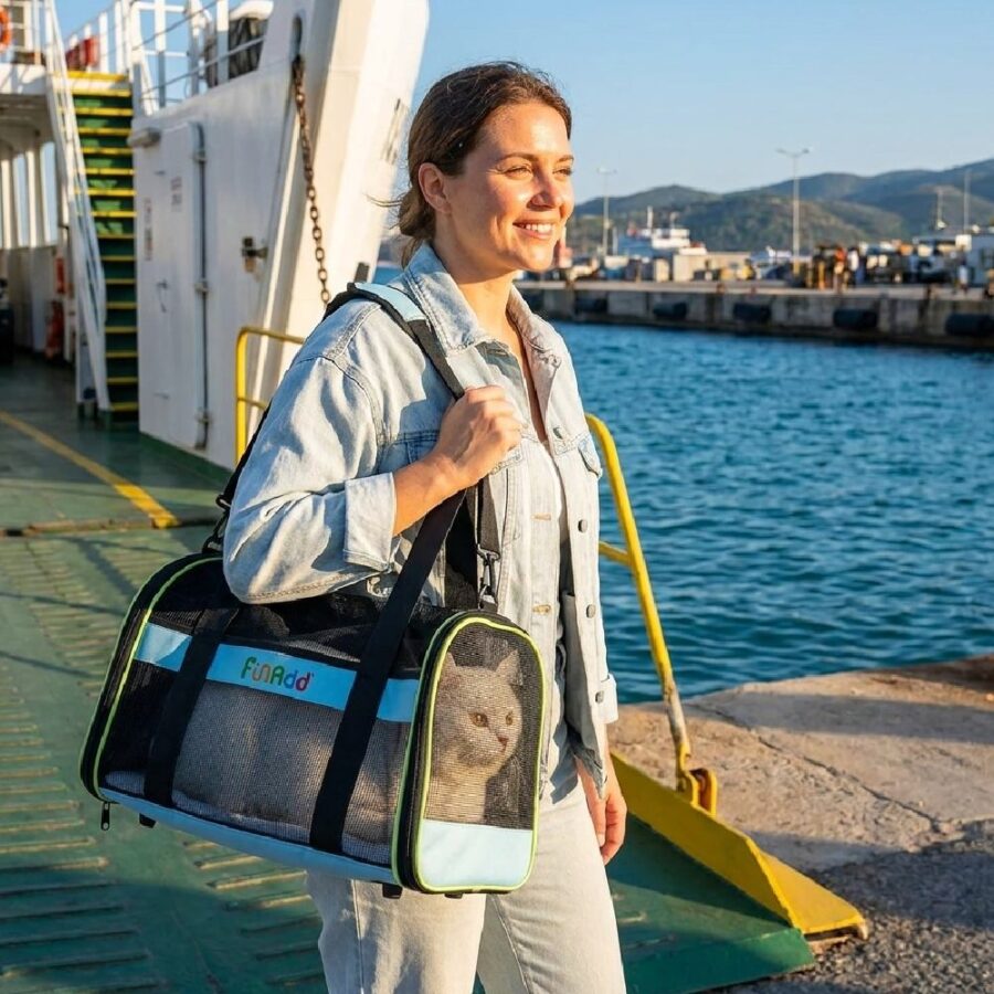 Femme souriante au bord de l'eau portant un sac vert et noir contenant un chat, sur un quai près d'un ferry